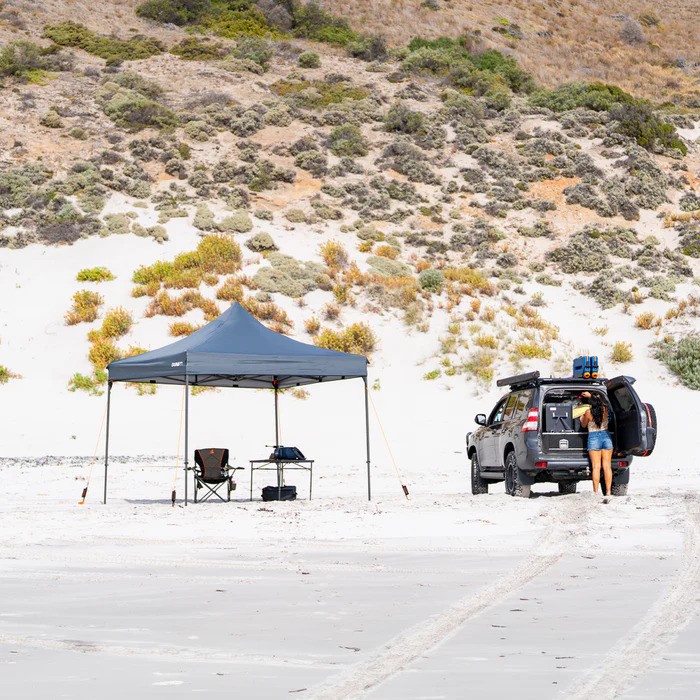 02 Tiegear Beach A woman works in her overland truck while her shade tent is secured into the sand with Tiegear equipment.
